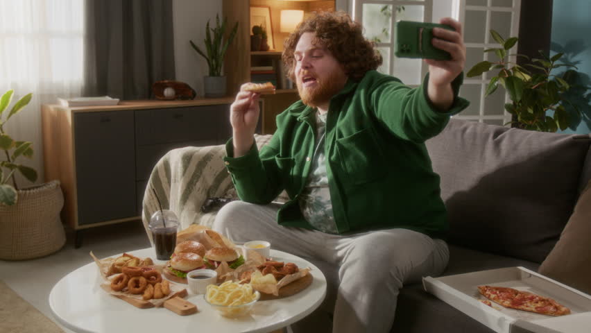 Young red-haired man talking about fast food on camera, holding smartphone in his hand and sitting on couch in his living room