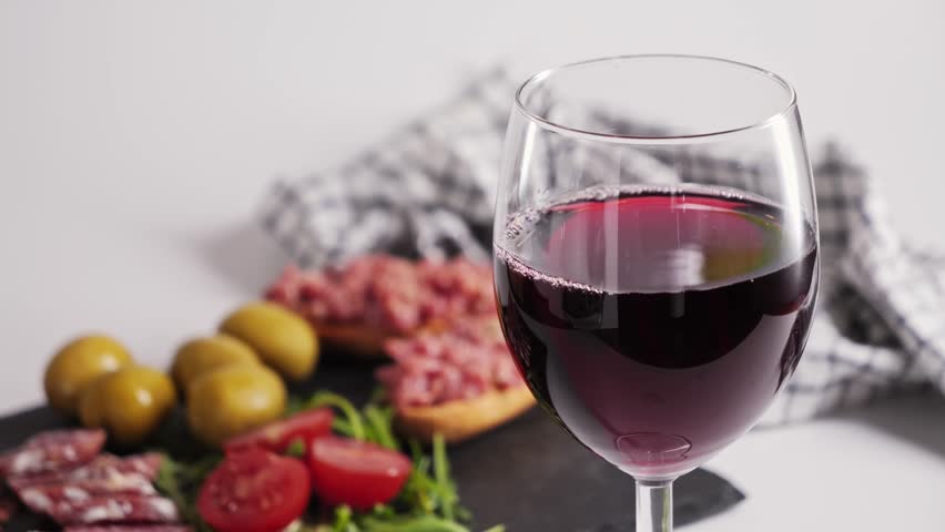 A glass of dry red wine on backdrop of traditional Spanish snacks such as fuet sausage, jamon and green olives. High quality 4k footage