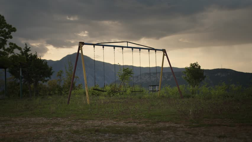 Static wide shot of an old, empty swingset in an overgrown clearing, with moody clouds and a mountain ridge silhouetted in the background.