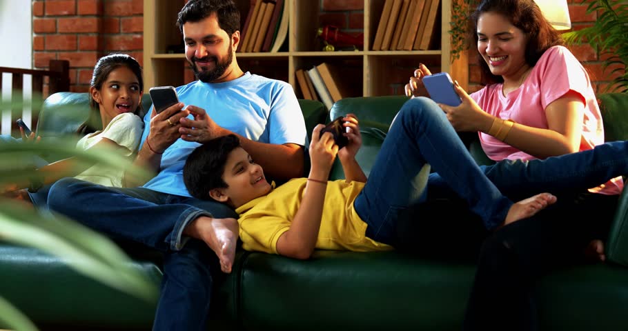 Indian family using smartphones individually while sitting together on living room couch, showing screen addiction in modern lifestyle, no interaction or bonding, lack of quality family time concept