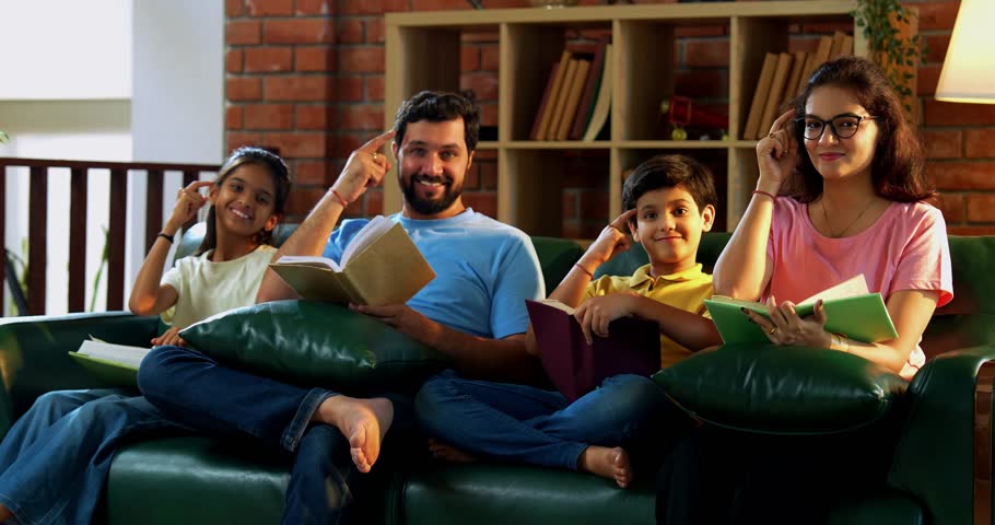 Family promoting book reading together while sitting on cozy couch at home, young Asian couple with children gesturing importance of developing reading habit for intelligence and self growth indoors