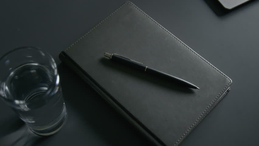 Closeup of sleek black notebook with pen placed on top, accompanied by clear glass of water on dark matte office desk