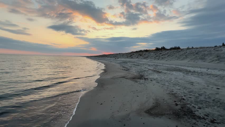 Tracking shot while calm waves gently lap the sandy shore at sunset.  Dunes covered in sea grass and shrubs line the coast of Graal-Müritz, Germany.  The sky is a mix of orange, pink, and blue.