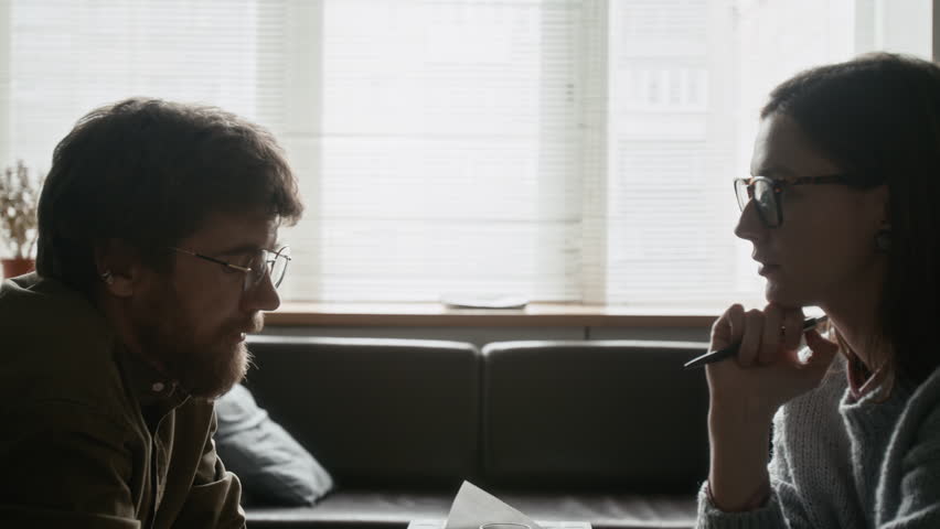 Side-view chest up shot of man and therapist sitting face to face, engaged in deep and focused conversation in softly lit counseling space
