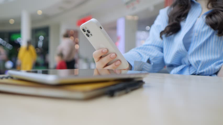 Woman is holding a cell phone and looking at it. She is sitting at a table with a stack of books and a laptop. The scene suggests that she might be working or studying, as she has multiple devices