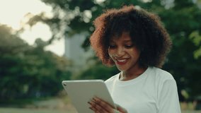 Portrait of positive African American female holding tablet computer in green summer park. Young woman watches visual content via digital device on city street. Urban style - Powered by Shutterstock - Get 15% off with code: PIKWIZARD15