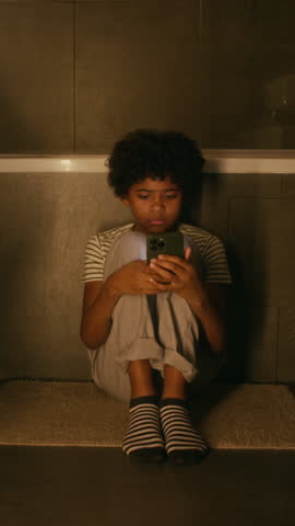 Full vertical of young black boy sitting alone on floor in dark bathroom, reading abusive messages on social media from bullies, crying and wiping off tears, suffering from loneliness and despair