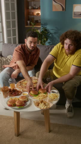 Vertical shot of two friends eating burgers and watching TV at home, having large fast food meal