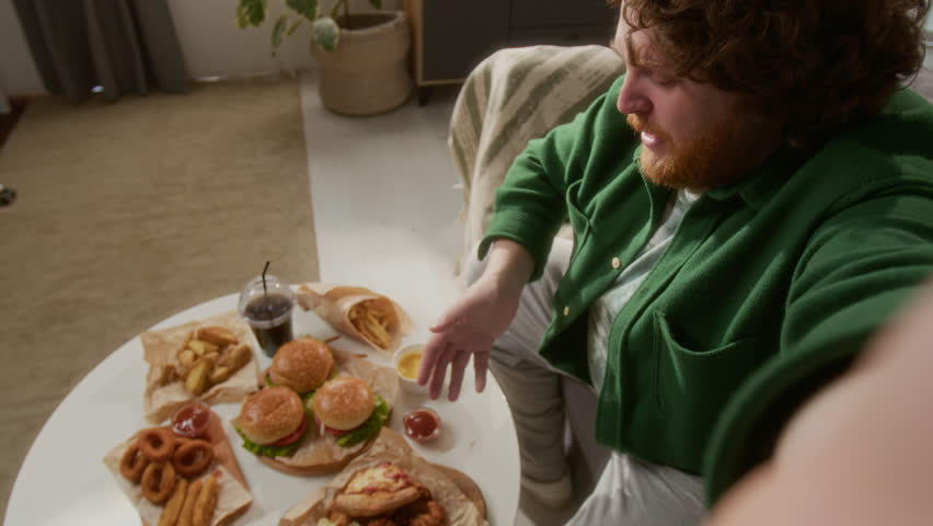 Bearded red-haired man filming food blog, talking to camera while eating fast food at home in living room