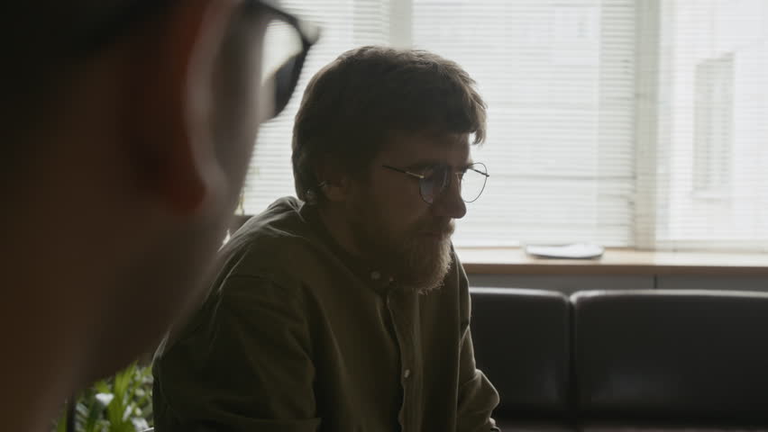 Young adult Caucasian man with beard sharing light moment with others during group therapy session, expressing openness and connection in supportive setting