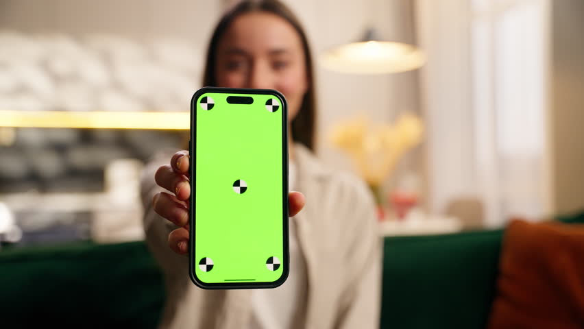 Happy Caucasian woman girl female user showing smartphone mobile phone with green screen relaxing on sofa living room home color chroma key template layout for advertising smiling looking camera app