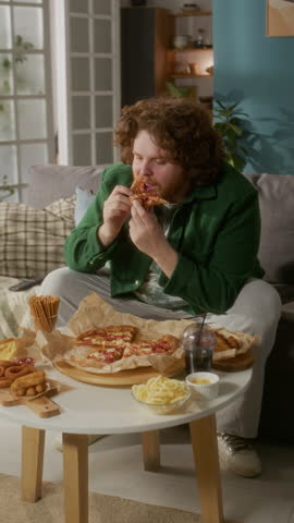 Vertical shot of chubby man hungrily eating pizza and other fast food snacks with soda at home in his living room
