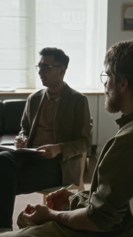 Vertical shot of Indian male therapist sharing warm, engaging smile while interacting with client during therapy session, creating welcoming and trusting atmosphere in modern office
