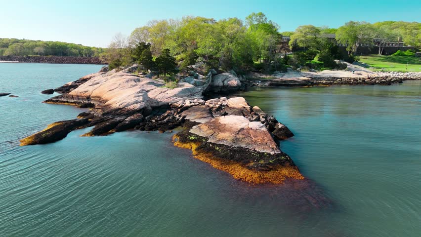 Aerial view of a rocky shoreline and small forested island surrounded by calm blue water. 