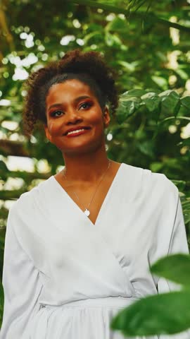 happy African American woman walking in tropical forest, hand stroking touching green palm tree leaves, smiling face girl enjoying summer nature tropics. White dress with sleeves. vertical video crop