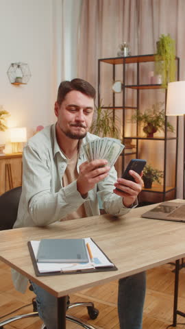 Planning family budget. Smiling Caucasian man counting money dollar cash use smartphone calculate domestic bills. Guy satisfied of income, saves salary money at home office table workplace. Vertical