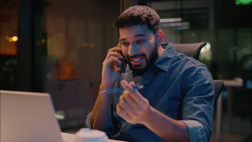 Indian stressed businessman furious business man talking mobile phone in night evening office displeased angry employer discuss problem laptop working overwork stress smartphone call arguing conflict