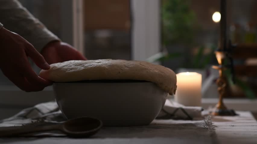 Close-up of man’s hands folding risen dough overflowing from bowl on table.