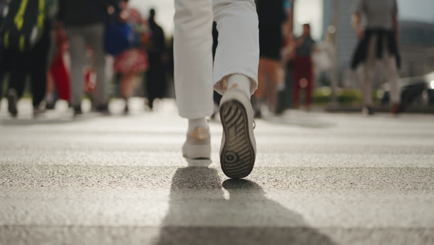 Back view following shot female feet steps walking pedestrian crossing street, sunny day, young woman wearing white pants and sneakers goes crosswalk. Low angle people legs stepping zebra asphalt