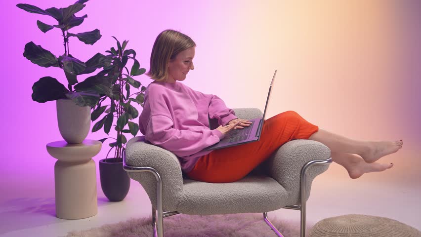 A cheerful woman types on her laptop while reclining barefoot in a cozy chair. Dressed in pink and orange, she enjoys a calm, colorful work moment in a modern space filled with soft textures