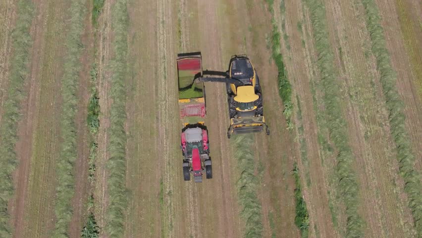 Slow-motion 70mm drone shot of a harvester and tractor with trailer working in sync from a front view, collecting and loading hay from neat rows on the ground during harvest operations.