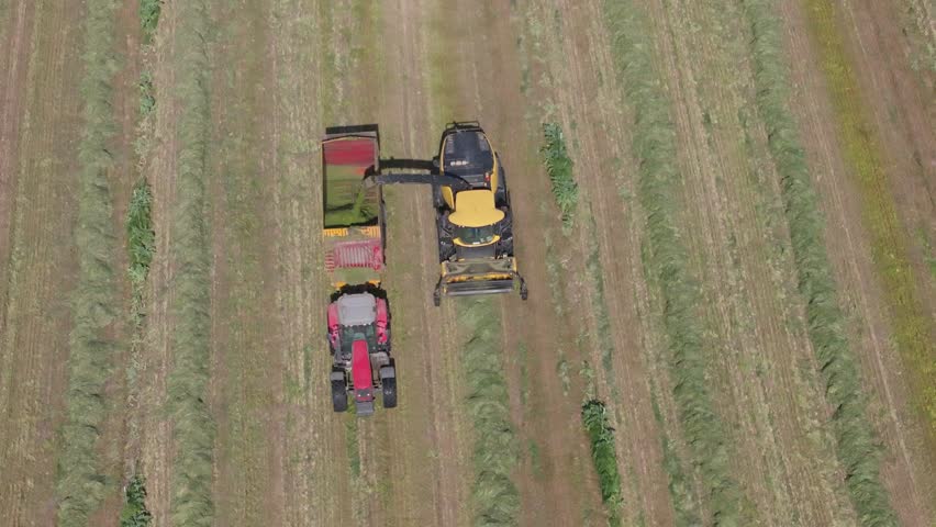 Slow-motion drone footage in 70mm showing a harvester collecting hay in rows and transferring it through a chute into a trailer pulled by a tractor driving alongside.