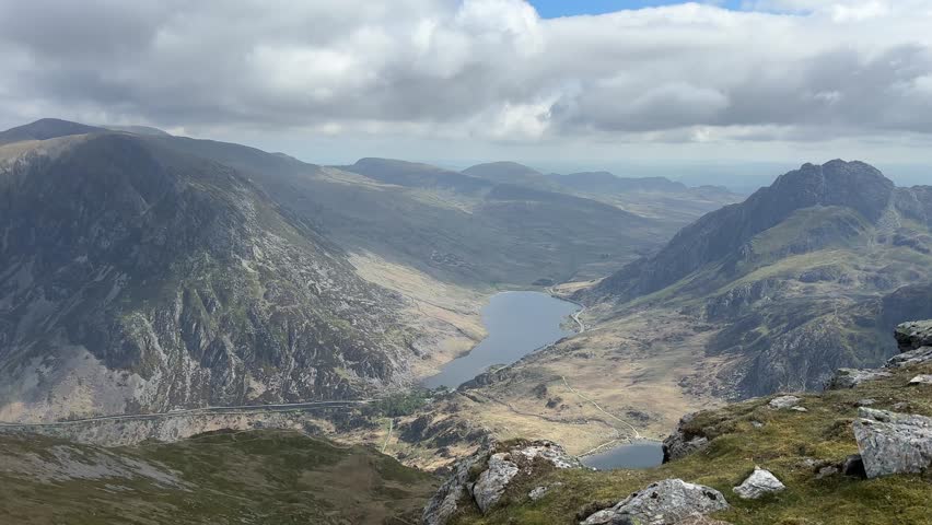 Scenic view from Y Garn summit in Snowdonia National Park showing mountain peaks, deep valley, rocky slopes and clear lake under partly cloudy sky, captured while hiking along trail.