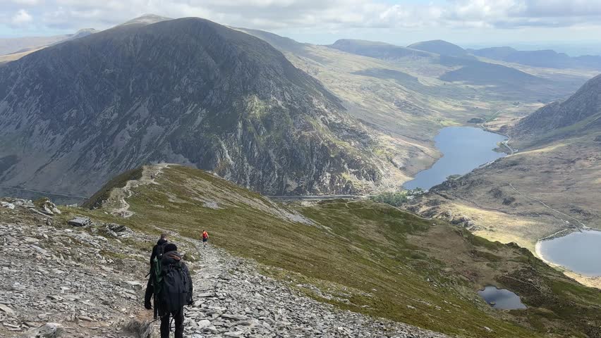 Hiker walking down rocky trail from Y Garn in Snowdonia National Park with wide view of mountain range, peaceful valley and blue lakes under cloudy sky during outdoor adventure in nature.