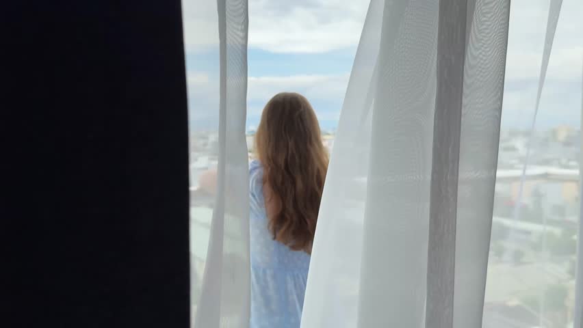 Girl standing on balcony, city visible behind tulle, rising on tiptoes in long blue dress, European woman resting in Asia, Vietnam