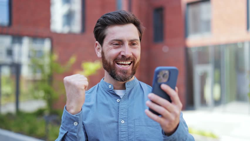 Successful lucky businessman in casual clothes expressing excitement with fist pump outdoors, celebrating success. Joyful man feeling enthusiastic and happy about good news on phone.