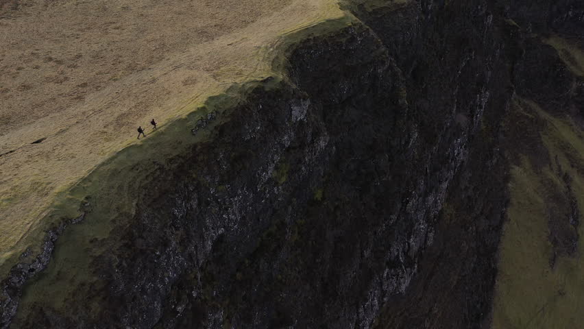 Drone pullback showing two hikers walking along a steep, barren cliff edge on the Trotternish Ridge, Isle of Skye, with exposed erosion and glacially carved landscape.