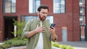 Portrait of ill man feels chest pain while reading bad news on his phone. He is standing outdoors and wearing casual clothes. Heart attack, health problem. Trouble breathing, health problems concept. - Powered by Shutterstock - Get 15% off with code: PIKWIZARD15