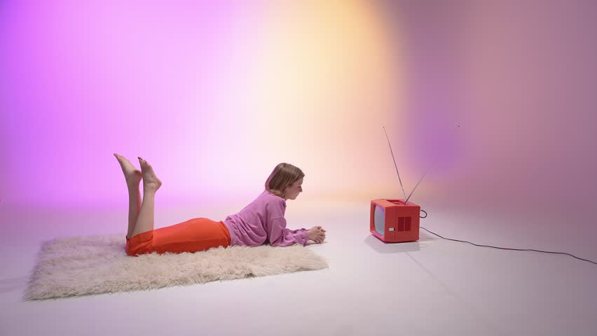 A young woman lies on a fluffy rug and reaches toward a vintage red television. The background glows with pink and yellow hues, blending nostalgic aesthetics with a cozy, retro-inspired vibe - Powered by Shutterstock - Get 15% off with code: PIKWIZARD15