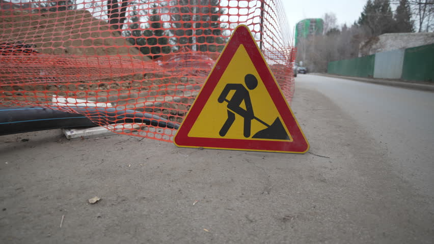 Rubber pipe winds across ground into hole by bright traffic sign Road Works. Indifferent pedestrians pass by in cold weather along sidewalk