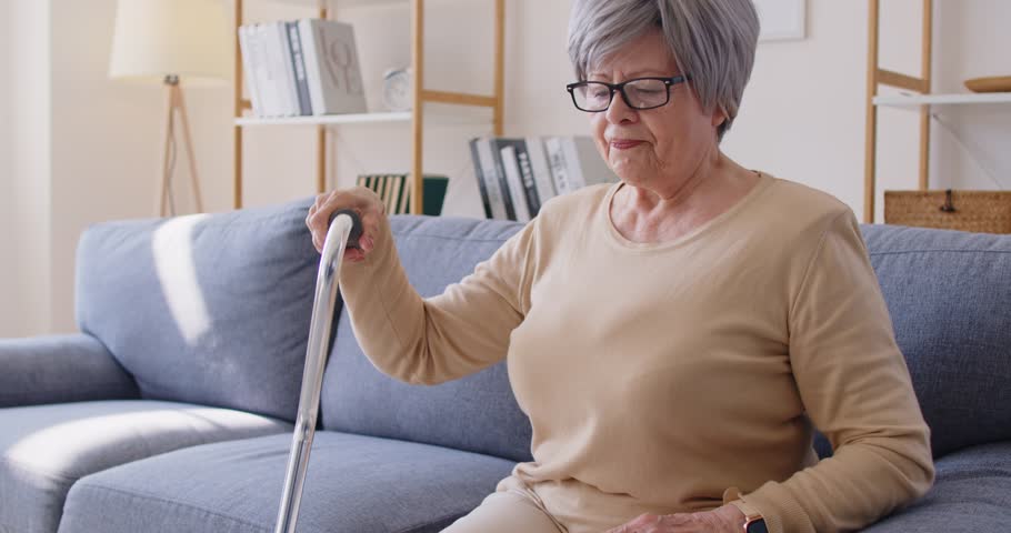 Elderly woman with gray hair using cane to get up from sofa in living room. Senior lady struggling with discomfort or joint pain, trying to stand up carefully and supporting herself with stick.