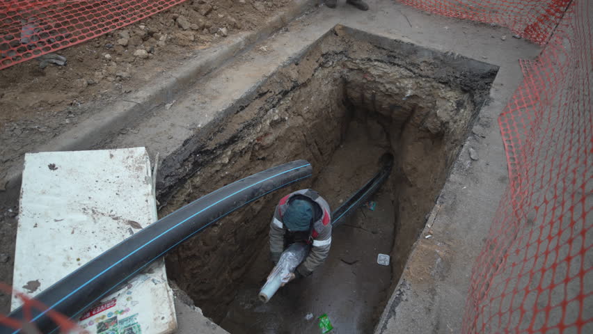 Sewer pit fenced with netting contains newly installed drainage machinery. Skilled worker carefully arranges system for optimal flow