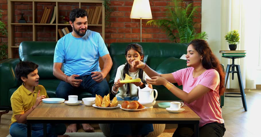 Indian family eating samosa snacks with tea at home, parents and children sitting in cozy living room enjoying fried food and warm drinks while chatting, laughing, and sharing quality bonding time