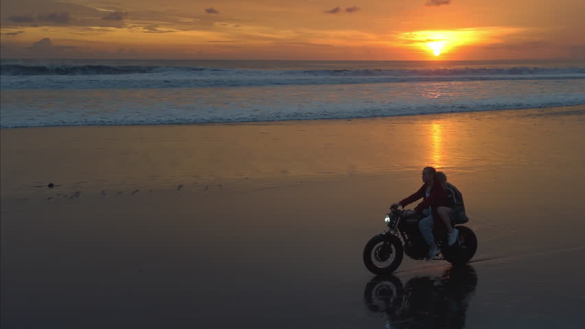 Couple rides along beach on motorcycle on a tropical island. Man and woman in road trip on a bike on the coast. Male and female ride a scooter on the ocean or sea shore. Bali Indonesia, sunset.