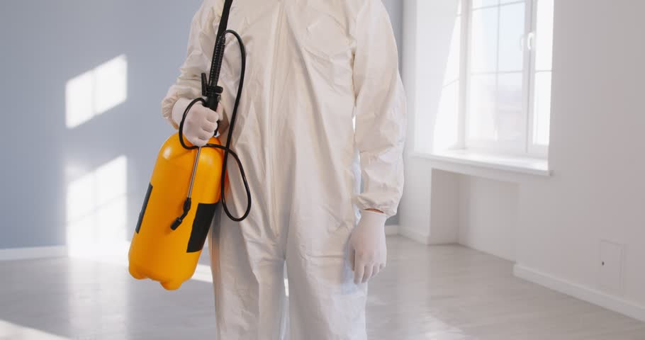 Portrait of pest control exterminator in white protective suit standing in empty room and smiling at camera. Professional contractor with yellow sprayer bottle putting off mask and gas respirator.