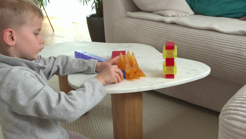 Young child engaged in creative play with colorful magnetic blocks on a table, constructing shapes with focus and curiosity. Indoor setting with cozy furniture in the background.