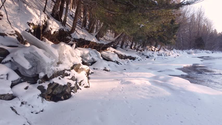 Snowy midday flyover of riverbank in Canada with winter forest and light mist