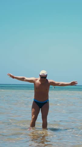 A man in swimming trunks, in sunglasses and a blazer is standing in shallow water and splashing , in the rays of the sun. he is sunbathing on a warm summer day, vacation at sea.