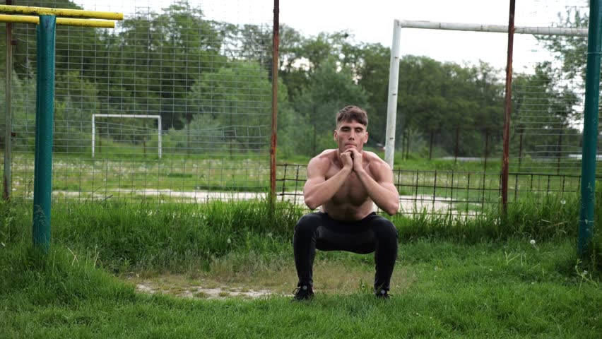 Male shirtless athlete doing squats at sports ground. Young sportsman working at summer nature. Strong sporty man exercising outdoor. Concept of active and sportive lifestyle. Slow motion