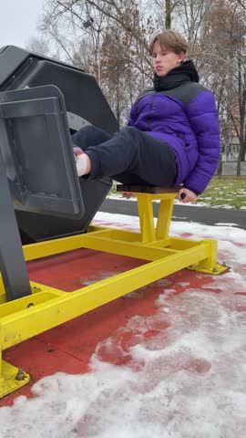 Teenager in a purple jacket is using a seated outdoor leg press machine in a snowy park setting while showcasing fitness activity