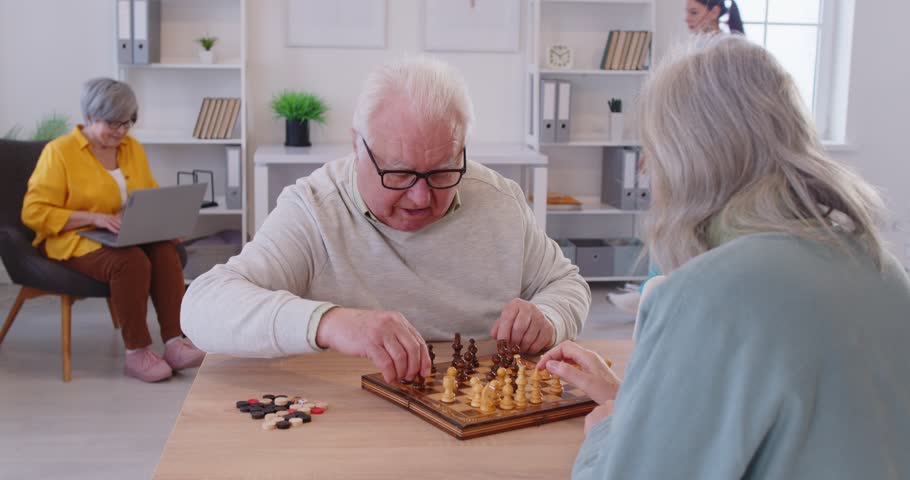 Happy elderly couple sitting at table and playing chess together in retirement home with senior woman using laptop. Cheerful pensioners having fun during leisure time, enjoying various activities.