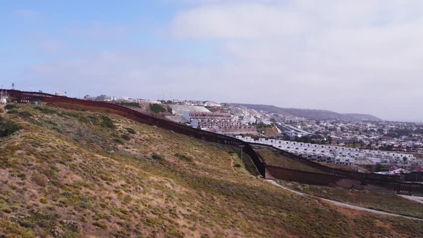 Drone pans over the hilly landscape where the U.S.–Mexico border fence splits the terrain and residential zones between Tijuana and San Diego.