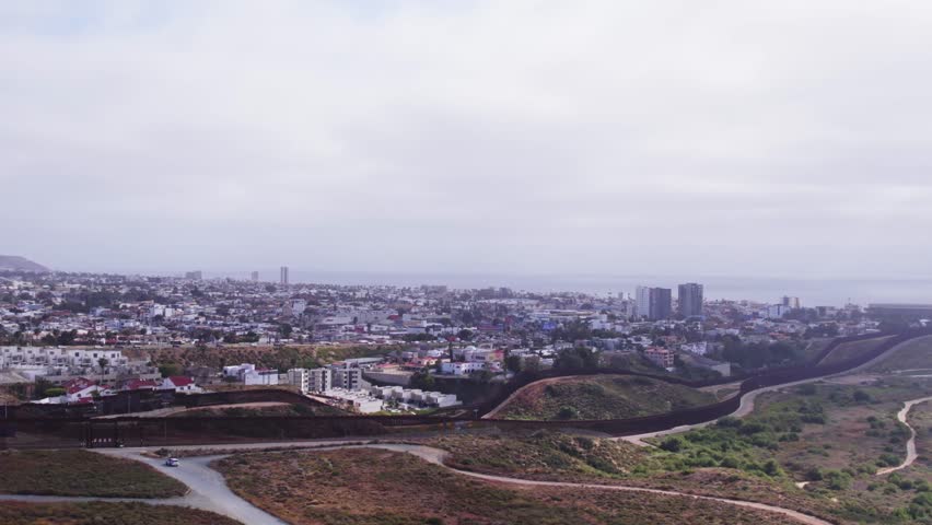Aerial view of the U.S.–Mexico border wall snaking through hills toward the cityscape of Tijuana under cloudy skies.