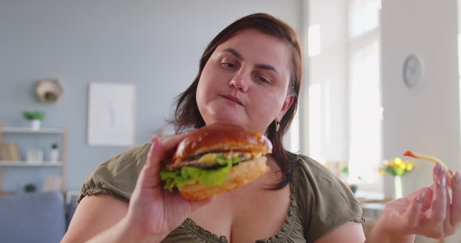 Close up of overweight fat woman enjoying big burger and french fries at home. Plus size female indulging in fast food, showing carefree attitude, love for tasty meal and unhealthy eating habits.