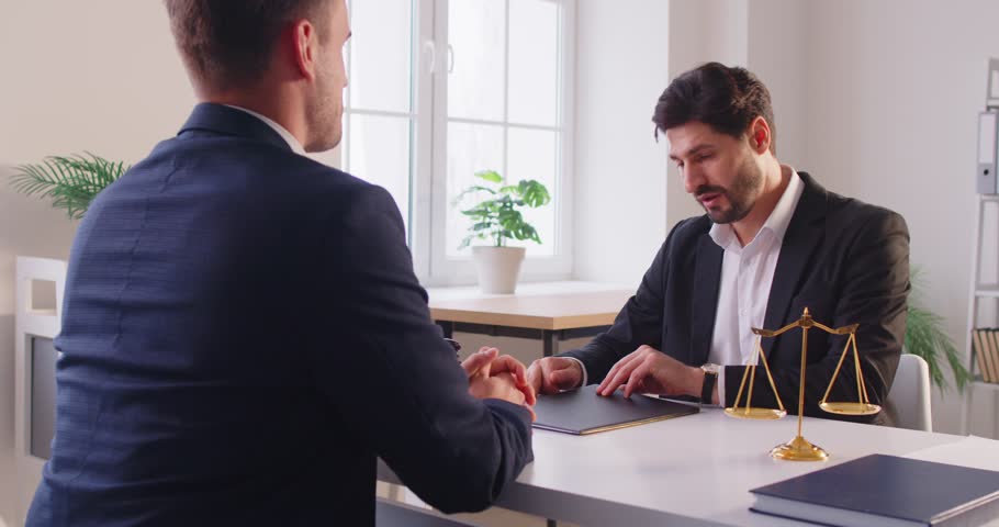 Professional male lawyer sitting at desk near judge scales with client, discussing legal documents or terms of contract. Attorney offering guidance and providing law services during consultation.