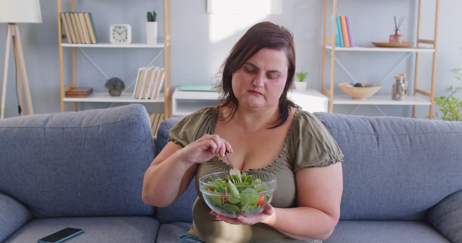 Unhappy fat plus-size woman reluctantly eats a vegetable salad at home living room. Her diet features healthy food choices, though she seems dissatisfied with the meal during leisure time.